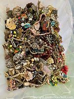 Photo showing a densely packed box with a large quantity of old, tangled, and damaged jewelry pieces including beads, chains, pendants, and brooch fragments.