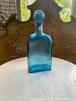 Large blue glass decanter with round stopper sitting on a table with a decorative background, showing front view