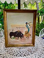 Full view of framed oil painting showing bullfighter and bull in a bullring under outdoor lighting, with greenery and white metal chairs in the background.