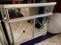 Front view of silver mirrored console table showing two doors with mirrored 'X' design panels and one drawer above with black knob.