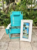 Photo showing the full turquoise Nautica folding beach chair with attached side pouch and the framed tropical print leaning against it on tiled outdoor ground with plants in the background.