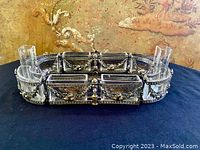 Front view showing entire silver ornate centerpiece with 4 rectangular glass containers and 2 taller candle glass inserts, placed on dark fabric with detailed wallpaper background.