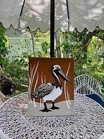 Front view of the framed Marüshka pelican textile art displayed outdoors on a white woven round table, showing the full image of the pelican with simple reeds on a two-tone background.