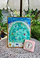 Photo of the giclee print on canvas showing a detailed, colorful pool with bathers in swimsuits, framed and standing on a white metal table next to a vintage pink clock.