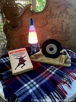Photo showing entire lot with wool blanket as background, lava lamp lit, chalkware record display holding a 45 record, and Art Buchwald book laying open.
