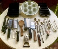 Wide shot of the lot showing knives, graters, wooden spatula, potato masher, forks, spoons, bottle stopper, corkscrew, and cast iron bakeware plates shaped like corn cobs.