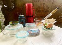 Overview of vintage and newer kitchen essentials on surface showing red Aladdin thermos, glass food storage containers with blue lids, colorful ceramic bowl, metal tin, measuring cup, ceramic cup with toothpicks.