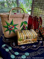 Overview of three vintage purses and three vintage belts arranged on a black surface outdoors, showing the woven basket handbag with floral applique, the red bucket purse, and the small illustrated handbag, along with the belt details.