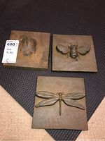 Three square metal art tiles with raised insect designs on a dark textured surface.