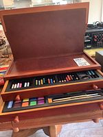 Open artists easel case showing watercolour pencils and coloured pencils arranged in a drawer with a pencil sharpener visible.