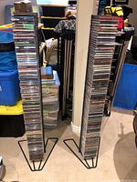 Two tall black metal CD towers filled with multiple music CDs, standing on carpet floor surrounded by storage bins and basement items.