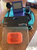 Purple Game Boy Advance SP open with visible buttons and screen, black charger with coiled cord, turquoise canvas case, and boxed orange GoPro Floaty on a wood table.
