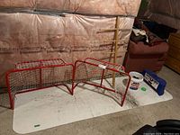 Overview photo showing two red kid size hockey nets, wooden rack, bucket of pucks, and plastic training mat on floor.