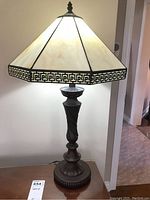 Front view of the metal and glass table lamp with geometric patterned stained glass shade showing detailed carved metal base and lamp lit.