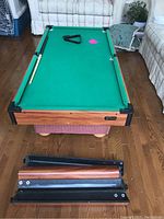 Top view of the Halex mini pool table assembled on one side, showing the green felt, table cues, and triangle rack on playing area. Detachable legs are placed beside the table.