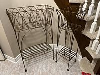 Pair of two metal plant holders with curved trellis design and wireframe base shelves, shown together in corner near staircase, on tiled floor