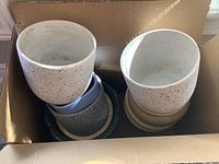 Box containing ceramic plant pots of various colors and sizes including white with speckles, gray, beige, and blue, plus matching saucers.
