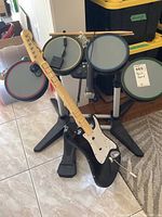 Front view of complete Rock Band drum kit and guitar controller set, including drum pads, foot pedal, microphone, drum sticks, and guitar controller placed on tiled floor.