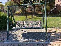 Front view of green metal swing set with cushioned seat on patio area