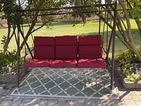 Metal outdoor swing set with red cushions. Shows full frame and bench setup with cushions on an outdoor patio with patterned rug.