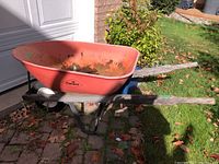Side and top view of orange Garant metal wheelbarrow with wooden handles in outdoor yard setting, showing wear and rust