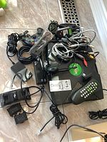 Photo showing original Xbox console with green logo, one wired controller, controller charger dock, headset, remote control, power adapter, and composite AV cables tangled and laid on floor.