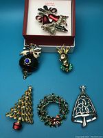 Photo showing six Christmas-themed brooches arranged on blue background, including bell, reindeer, trees, wreath, and bow, some in open box with Valerie Stevens branding