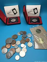 Overview of two 1970 Canadian silver dollars in red clamshell cases alongside a sealed package containing two Coca-Cola Olympic coins with a spread of various world coins in front on blue background.