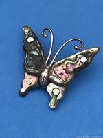 Front view showing colorful abalone shell inlaid butterfly wings, sterling silver body and curled antennae on a blue background.