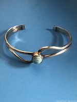 Front angled view of sterling silver cuff bracelet with turquoise-colored round stone centerpiece on a blue background.