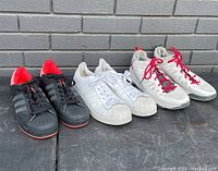 Overview of three pairs of men's sneakers set on pavement: black Adidas Superstars with red details, white Adidas Superstars, and white Nike Air Jordans with red laces.