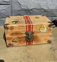 Front view of the antique doll trunk showing travel stickers, metal latch closure, and red stripes on brown exterior.