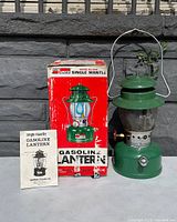 Photo of the green Ash Flash gasoline lantern model 1022, original box, and instruction manual placed outdoors against stone wall.