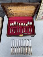 Photo of partial set of forks and spoons arranged in original wooden box with red velvet interior, knives laid out in front