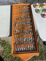 Full front view of the oak wood display rack filled with collector souvenir spoons with a glass front cover, set outdoors on pavement.