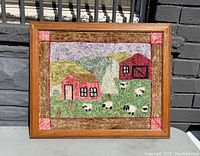 Front view of vintage hooked rug in wood frame showing country landscape with small houses and grazing sheep.