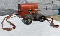 Binoculars and original brown leather carrying case with strap displayed outdoors on concrete surface.