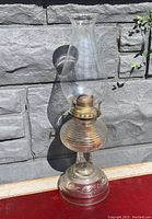 Full view of vintage clear glass oil lamp with hobnail chimney placed against stone wall background.