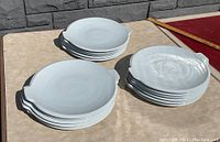 Sets of thick, heavy white porcelain chargers arranged in three stacks on a table outdoors, showing the spout-like extension on the rim