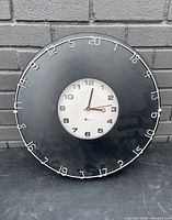 Full view of the round black wall clock. Clock face is centered with black and silver numbers. Outer ring has white wire numerical markings 1-24. Displayed against grey brick background.