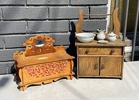 Photo of two vintage wooden dollhouse furniture pieces: a carved trunk with pink fabric lining and a small mirror, and a washstand with a drawer, cabinet, and ceramic wash set on top.