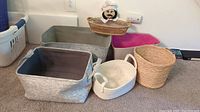 Six assorted baskets of different sizes and materials arranged on carpeted floor against a white wall.