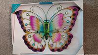 Photo of the butterfly wall ornament in its box showing vibrant wings with multicolors and decorative circular elements.