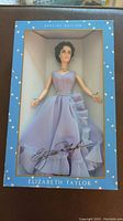 Photo of Elizabeth Taylor doll wearing lavender ruffled gown, inside unopened box with clear front displaying the doll and signature.