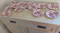 Twelve fine china plates with red floral and pastoral scene pattern spread on wooden drawer surface