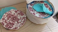 Set of 12 octagonal medium-sized plates with red floral and bird design, some visible stacked inside a zippered round holder with blue tissue paper inside.