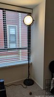 Full view of the floor lamp powered on, showing the round lamp head and black stand next to a window with blinds.