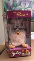 Front view of electronic Furby toy in original purple box with clear front window showing white-fur Furby with plastic glasses