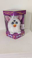 Front view of white Furby toy inside its original box with purple base and clear plastic window showing the Furby.