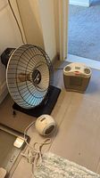 Three electric heaters placed on floor; one infrared heater with metal cage, one compact ceramic heater, and one small spherical heater.